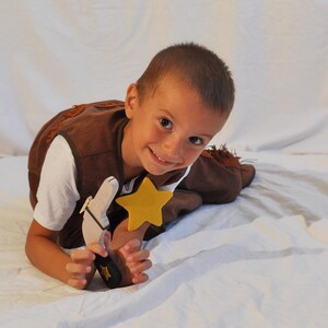 May include: A brown vest with fringe details, a white t-shirt, a wooden toy pistol with a black handle, and a yellow felt star badge. The vest features a dark brown trim and the toy pistol has a small yellow star emblem on the handle grip.