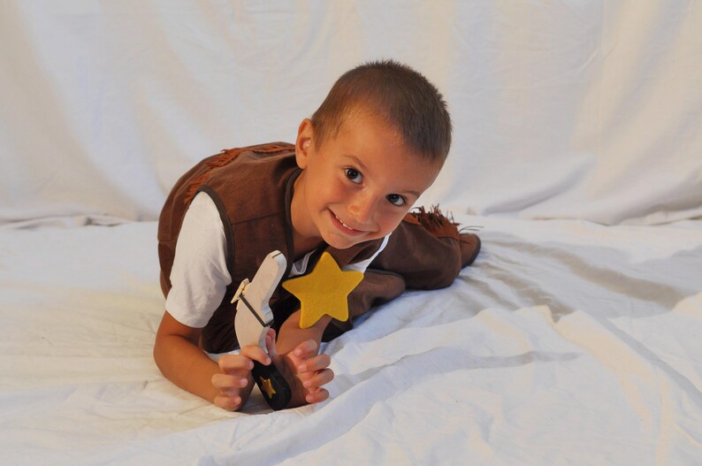 May include: A brown vest with fringe details, a white t-shirt, a wooden toy pistol with a black handle, and a yellow felt star badge. The vest features a dark brown trim and the toy pistol has a small yellow star emblem on the handle grip.