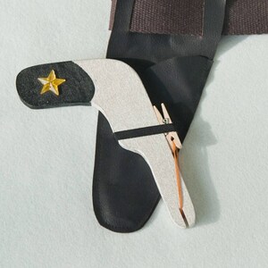 May include: A black leather holster featuring a silver-toned, boomerang-shaped tool with a black tip and a yellow star emblem. The tool is secured with a small wooden clothespin and a thin black strap, resting against the dark leather holster surface.