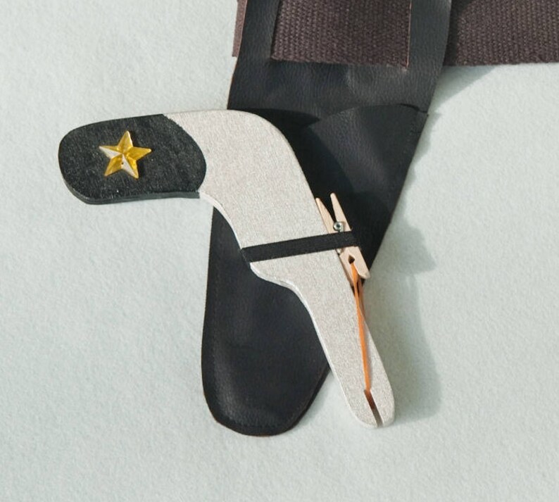 May include: A black leather holster featuring a silver-toned, boomerang-shaped tool with a black tip and a yellow star emblem. The tool is secured with a small wooden clothespin and a thin black strap, resting against the dark leather holster surface.