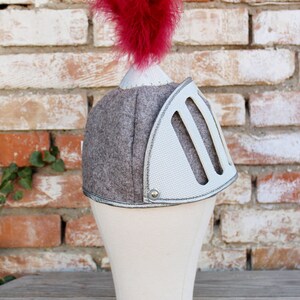 May include: A gray and white knight helmet with a red feather on top. The helmet is on a white mannequin head.