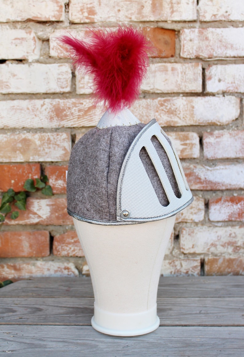 May include: A gray and white knight helmet with a red feather on top. The helmet is on a white mannequin head.