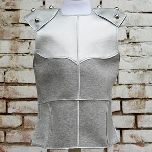 Puede incluir: Una blusa sin mangas de color plateado y gris con ribetes plateados y tachuelas plateadas en los hombros.