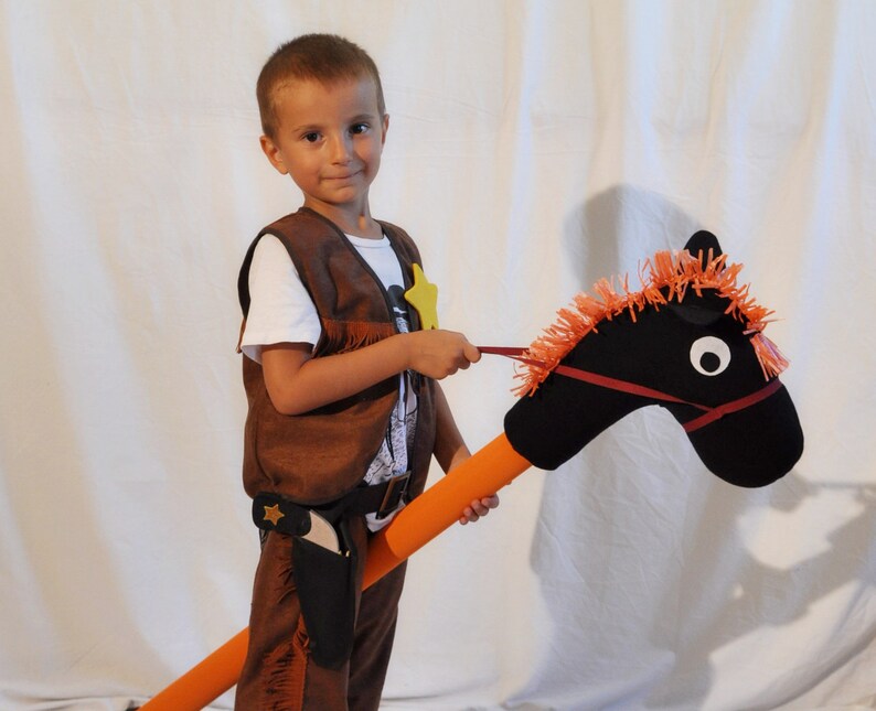 May include: A hobby horse with a black fabric head, orange yarn mane, and a long orange foam pole. The horse features a red bridle and white circular eyes. It is paired with a brown vest and pants set, including a holster and a yellow star badge.