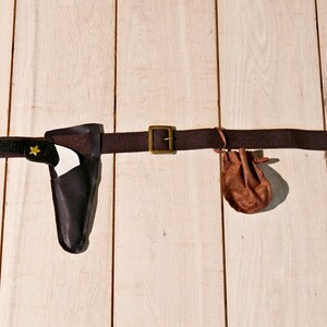 May include: A dark brown fabric belt featuring a brass-toned metal buckle, a black holster containing a white toy pistol with a small yellow star emblem, and a small brown drawstring pouch attached to the side of the belt strap.