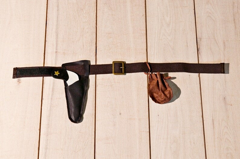 May include: A dark brown fabric belt featuring a brass-toned metal buckle, a black holster containing a white toy pistol with a small yellow star emblem, and a small brown drawstring pouch attached to the side of the belt strap.