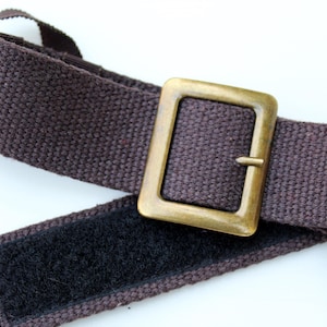 Pode incluir: Um cinto de tecido castanho escuro com uma fivela retangular de metal em tom de lat&atilde;o envelhecido. O cinto inclui uma tira de fecho de velcro preto no lado interior e uma presilha de fita castanha para prender a extremidade da correia.
