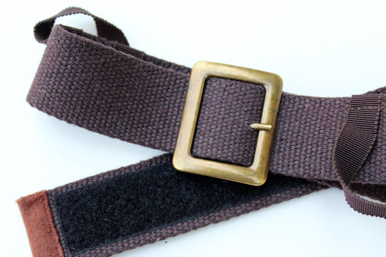 Pode incluir: Um cinto de tecido castanho escuro com uma fivela retangular de metal em tom de lat&atilde;o envelhecido. O cinto inclui uma tira de fecho de velcro preto no lado interior e uma presilha de fita castanha para prender a extremidade da correia.