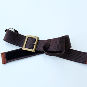 Pode incluir: Um cinto de tecido castanho escuro com uma fivela met&aacute;lica retangular em tom de lat&atilde;o. O cinto inclui uma tira de fecho de velcro preto no lado interior e possui extremidades revestidas com tecido castanho para um acabamento cuidado.