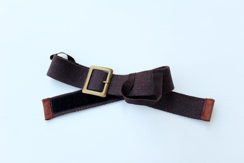 Pode incluir: Um cinto de tecido castanho escuro com uma fivela met&aacute;lica retangular em tom de lat&atilde;o. O cinto inclui uma tira de fecho de velcro preto no lado interior e possui extremidades revestidas com tecido castanho para um acabamento cuidado.
