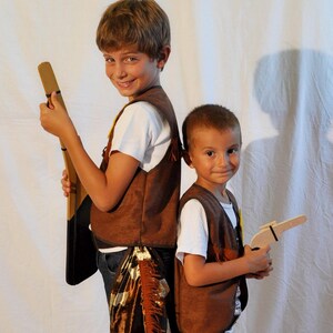 May include: Two brown suede-like vests with fringe detailing, worn over white t-shirts. One vest is paired with brown and white cow-print chaps featuring fringe. The accessories include a wooden toy rifle and a wooden toy pistol held by the wearers.