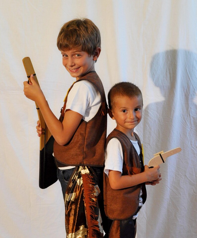 May include: Two brown suede-like vests with fringe detailing, worn over white t-shirts. One vest is paired with brown and white cow-print chaps featuring fringe. The accessories include a wooden toy rifle and a wooden toy pistol held by the wearers.