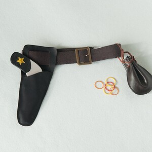 May include: A brown fabric belt with a brass-toned buckle, featuring a black leather holster containing a toy pistol with a yellow star emblem, a small dark brown leather pouch attached to the side, and several small orange and yellow rubber bands.