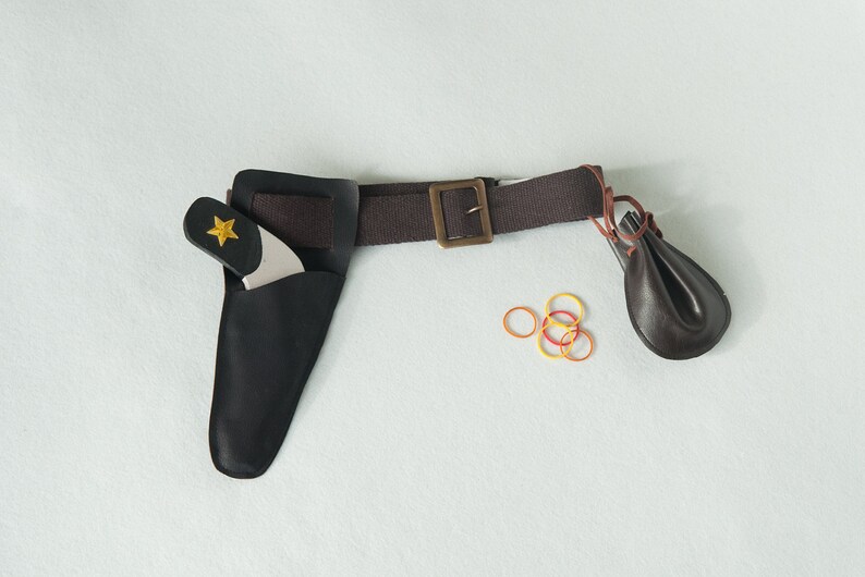May include: A brown fabric belt with a brass-toned buckle, featuring a black leather holster containing a toy pistol with a yellow star emblem, a small dark brown leather pouch attached to the side, and several small orange and yellow rubber bands.