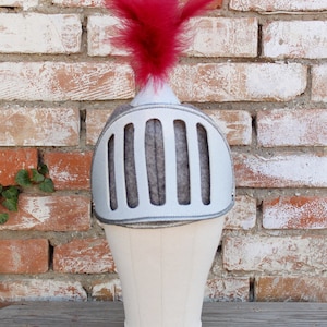 May include: A white knight helmet with a red feather on top. The helmet has a gray mesh face guard and is on a white stand.