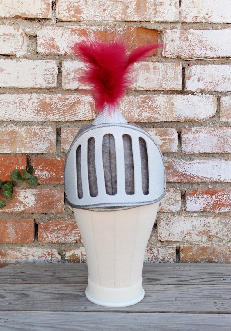 May include: A white knight helmet with a red feather on top. The helmet has a gray mesh face guard and is on a white stand.