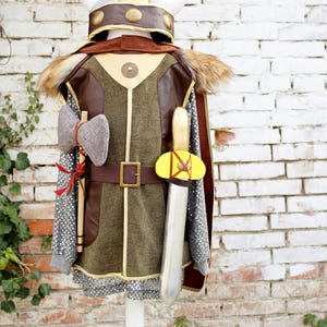 Pode incluir: Um conjunto de fantasia com um capacete com chifres, uma t&uacute;nica castanha e verde-azeitona com detalhes dourados, uma capa de pele sint&eacute;tica, uma camisa cinzenta estilo cota de malha, um machado cinzento e uma espada prateada com punho amarelo.