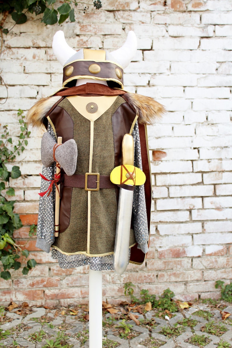 Pode incluir: Um conjunto de fantasia com um capacete com chifres, uma t&uacute;nica castanha e verde-azeitona com detalhes dourados, uma capa de pele sint&eacute;tica, uma camisa cinzenta estilo cota de malha, um machado cinzento e uma espada prateada com punho amarelo.