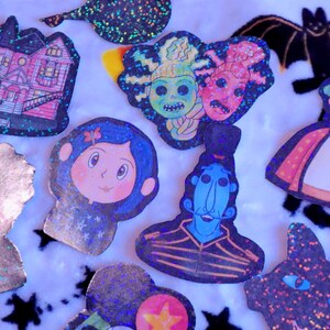 May include: A collection of nine glittery iron-on patches featuring various characters and designs, including a house, a girl with a blue bow, a blue creature with a mustache, a woman with glasses, and a black cat.