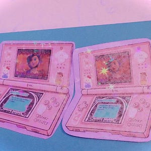 Op de afbeelding: Twee roze en witte Nintendo DS-gameconsole stickers met een cartoon afbeelding van een meisje op het scherm. De stickers hebben de tekst "START" en "SELECT" onderaan.