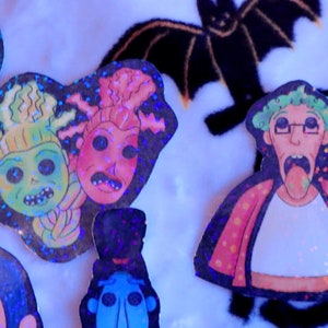 May include: Five holographic stickers featuring cartoon characters with exaggerated features. The characters are green, pink, blue, and white with black outlines. The stickers are on a white fuzzy surface.