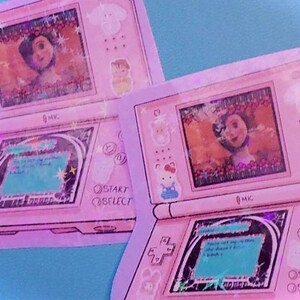 Op de afbeelding: Twee roze handheld gaming-apparaten met een cartoonfiguur op het scherm. De apparaten zijn roze en wit met een zwarte omtrek. Het scherm toont een cartoonfiguur tegen een blauwe achtergrond. De apparaten hebben knoppen met de tekst "Start" en "Select".