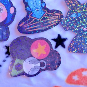 May include: A set of five holographic stickers featuring characters from the movie "Coraline". The stickers are on a white fuzzy background. The stickers are of Coraline, the Other Mother, the Cat, and two other characters.