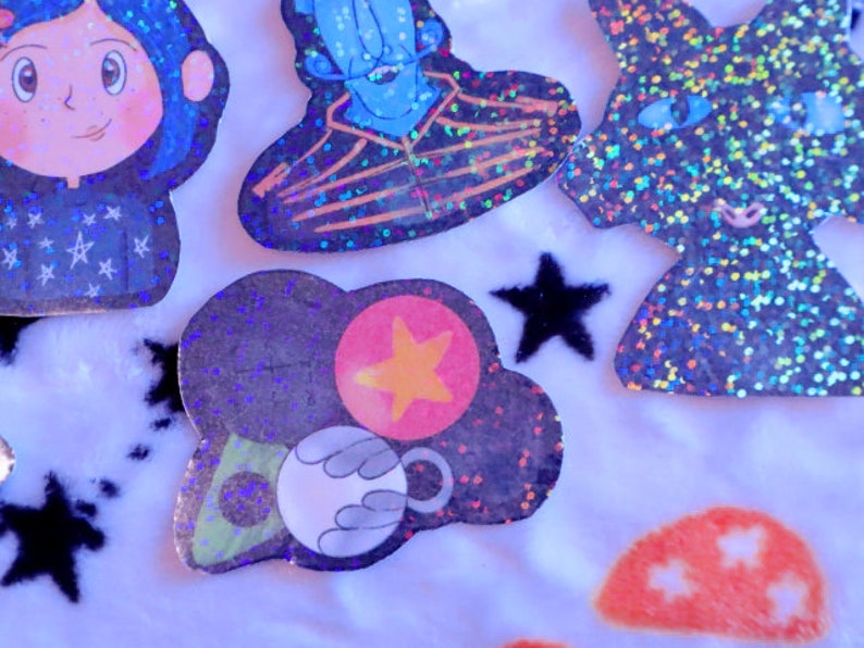 May include: A set of five holographic stickers featuring characters from the movie "Coraline". The stickers are on a white fuzzy background. The stickers are of Coraline, the Other Mother, the Cat, and two other characters.