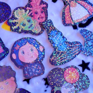 May include: A collection of colorful, glittery stickers featuring various characters and designs. The stickers are on a white background and include a house, a girl with blue hair, a monster, a witch, and a star.