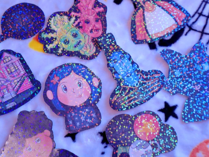 May include: A collection of colorful, glittery stickers featuring various characters and designs. The stickers are on a white background and include a house, a girl with blue hair, a monster, a witch, and a star.