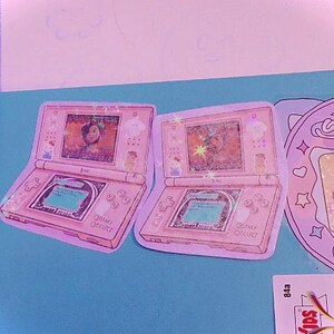 Op de afbeelding: Twee roze Nintendo DS handheld-spelconsoles met een scherm waarop een cartoonfiguur te zien is. De consoles bevinden zich op een lichtblauwe achtergrond.