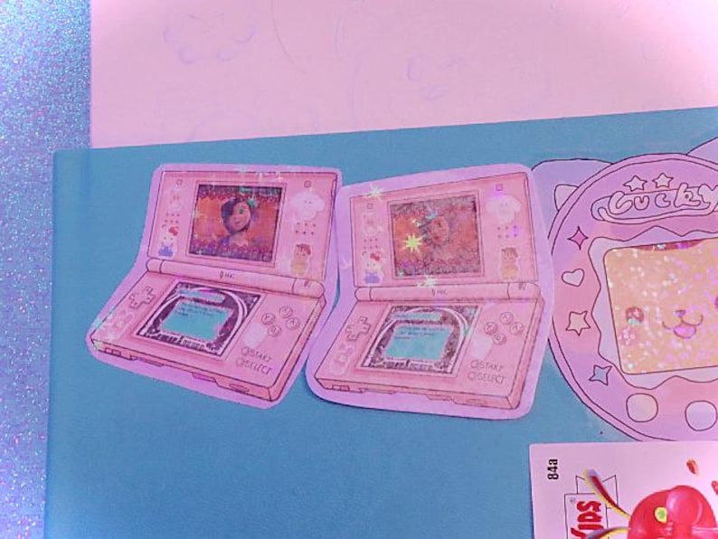 Op de afbeelding: Twee roze Nintendo DS handheld-spelconsoles met een scherm waarop een cartoonfiguur te zien is. De consoles bevinden zich op een lichtblauwe achtergrond.