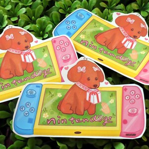 May include: Stickers featuring a cartoon dog on a video game console. The console has a yellow screen with a green background and the text "Nintendogs". The dog is brown with a pink bow and scarf. The console's controllers are blue and pink.