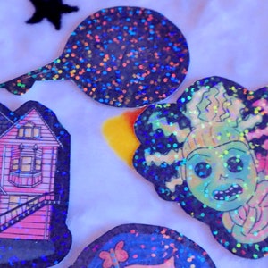 May include: A set of four holographic stickers featuring a pink house, a green monster with a toothy grin, a pink monster with a tongue sticking out, and a black and silver glitter circle.