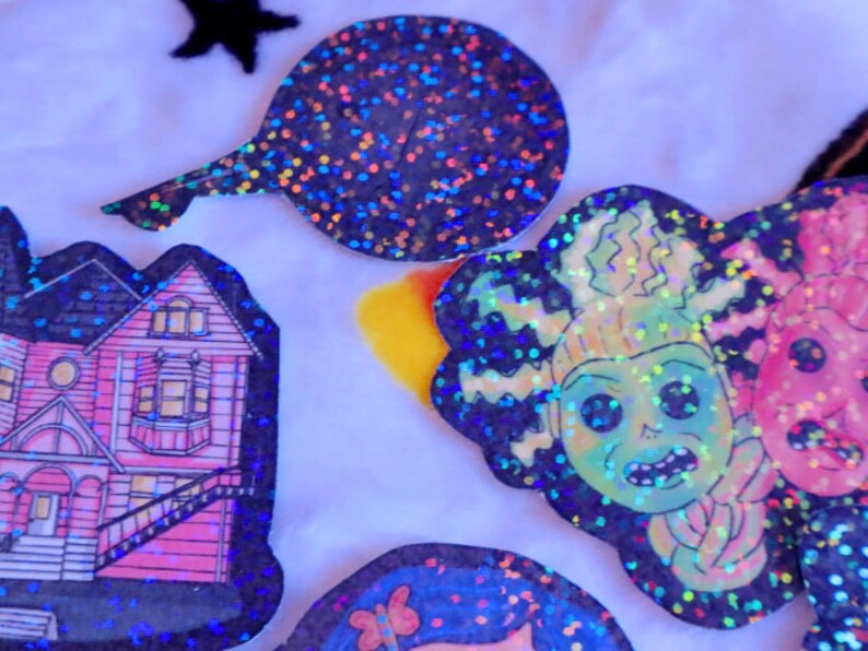May include: A set of four holographic stickers featuring a pink house, a green monster with a toothy grin, a pink monster with a tongue sticking out, and a black and silver glitter circle.