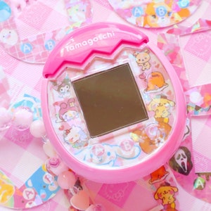 May include: Pink Tamagotchi toy with a screen and various cartoon characters on the front. The toy is surrounded by pink and white decorative stickers.