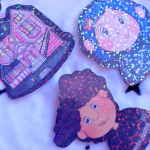 May include: Three glitter-covered stickers featuring a pink house, a girl with blue hair, and a boy with black hair. The stickers are on a white fuzzy surface.