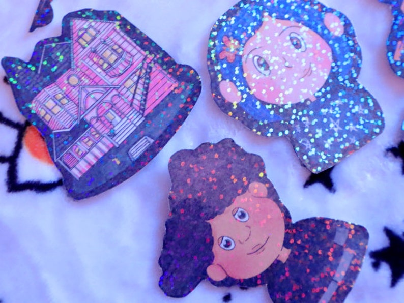 May include: Three glitter-covered stickers featuring a pink house, a girl with blue hair, and a boy with black hair. The stickers are on a white fuzzy surface.