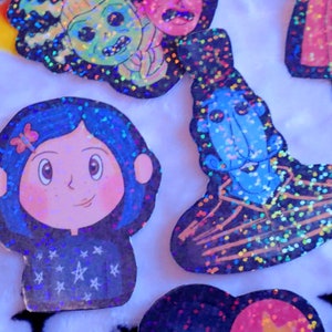 May include: A set of colorful, glittery stickers featuring characters from the movie "Coraline". The stickers feature Coraline, the Other Mother, and the cat. The stickers are perfect for adding a touch of whimsy to your laptop, water bottle, or planner.