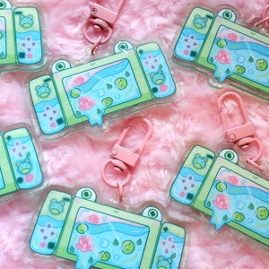 May include: A set of six green and blue frog-themed keychains with a cartoon video game console design. Each keychain features a clear acrylic background with a pink and green frog design. The keychains have a pink metal clasp.