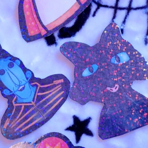 May include: A set of four holographic glitter stickers featuring a blue character with a top hat, a black cat, a red and white rocket, and a black star. The stickers are on a white fuzzy background.