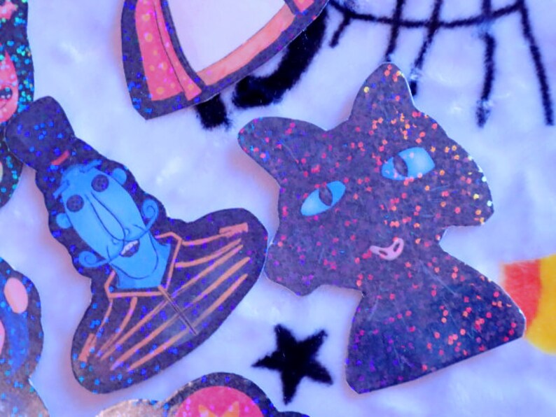 May include: A set of four holographic glitter stickers featuring a blue character with a top hat, a black cat, a red and white rocket, and a black star. The stickers are on a white fuzzy background.