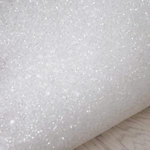 May include: Close-up of white glitter fabric. The fabric has a soft texture and is covered in small, sparkling glitter particles. The glitter catches the light, creating a shimmering effect. The fabric is folded over, revealing its thickness.