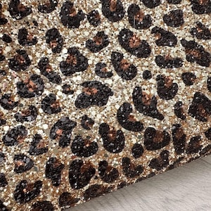 Puede incluir: Primer plano de una superficie brillante con un estampado de leopardo. El diseño presenta manchas marrones oscuras y negras sobre un fondo de purpurina dorada y plateada. La textura es brillante y llamativa, adecuada para diversos proyectos de manualidades.