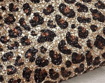 May include: Close-up of a shimmering surface with a leopard print pattern. The design features dark brown and black spots on a gold and silver glitter background. The texture is sparkly and eye-catching, suitable for various craft projects.