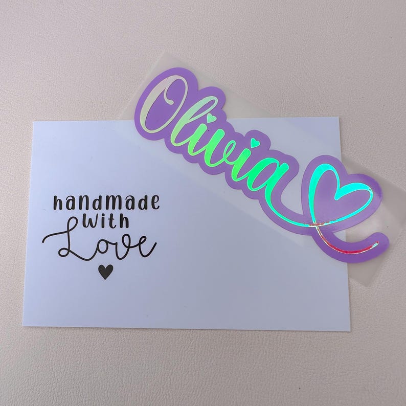 May include: A white card with the text "handmade with Love" and a heart. A purple and green holographic sticker with the name "Olivia" and a heart design is placed on the card.