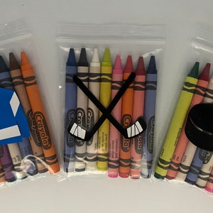 May include: Three clear plastic bags filled with Crayola crayons. The bags are decorated with hockey-themed cutouts. One bag has a hockey stick cutout, another has a hockey puck cutout, and the third has a hockey jersey cutout.