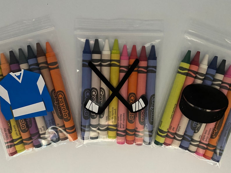 May include: Three clear plastic bags filled with Crayola crayons. The bags are decorated with hockey-themed cutouts. One bag has a hockey stick cutout, another has a hockey puck cutout, and the third has a hockey jersey cutout.
