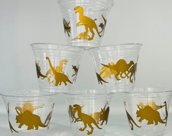 May include: Six clear plastic cups with gold dinosaur silhouettes. The dinosaurs include a T-Rex, Triceratops, Stegosaurus, and others.