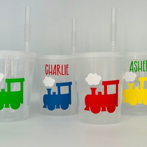 May include: Four clear plastic cups with straws and personalized names. Each cup features a colorful train design with a green, blue, red, and yellow train. The names on the cups are Charlie, Asher, and two other names that are not visible.